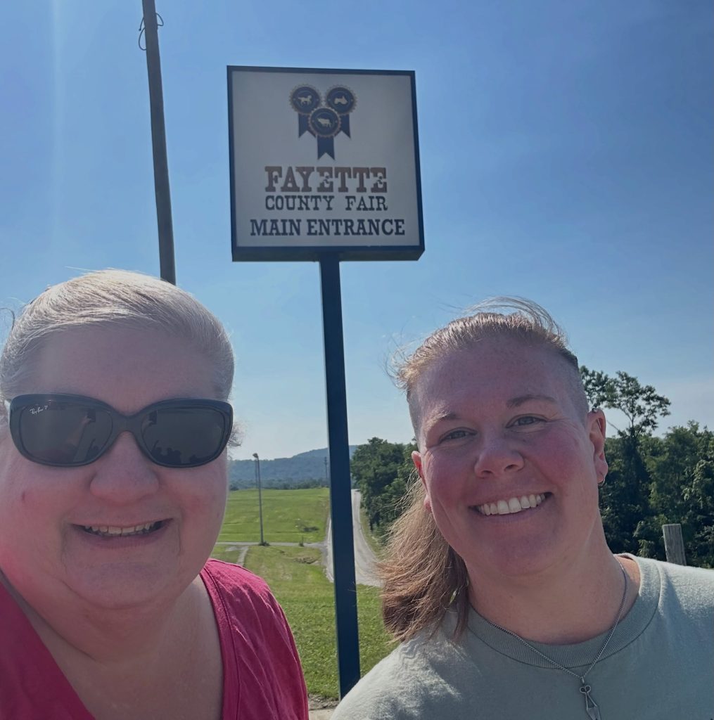 Two women smiling at Fayette County Fair main entrance sign on a sunny day.